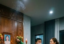 Trên bức tường lớn đối diện sofa, một di ảnh được treo trang trọng. Khung gỗ đen, vòng hoa trắng, nhang còn đang cháy dở.