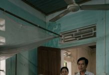 Hưng đứng trước cửa phòng mẹ, nhưng không bước vào. Anh chỉ nhìn vào trong khoảng ba giây rồi quay đi – vẻ mặt hơi buồn.